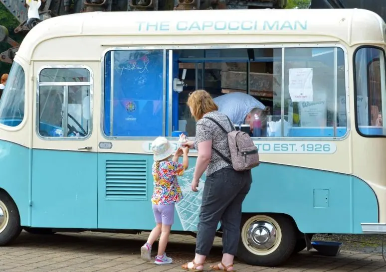 Woman and child ordering from a vintage cream and blue ice cream van at an outdoor venue.