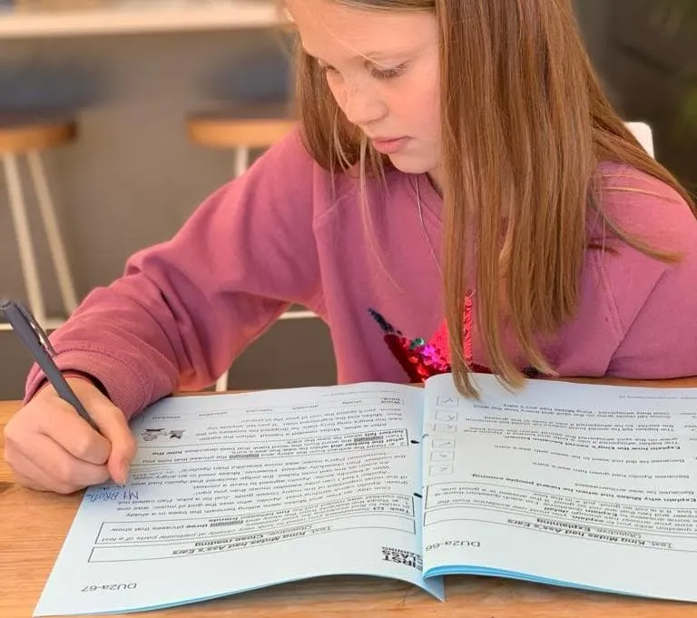 Child in pink top concentrating while writing in an open workbook at a wooden table.