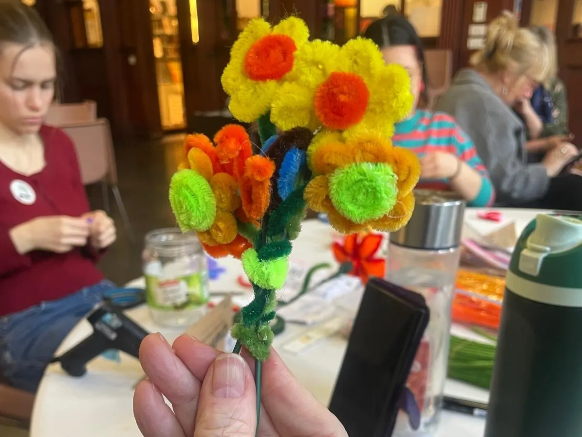 Hand holding a colourful pompom flower craft during a workshop with participants seated at a table.