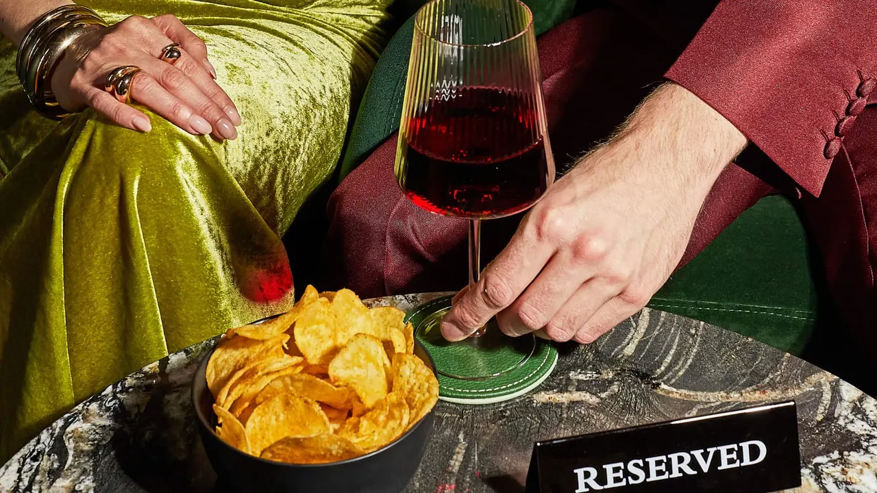 Two people in colourful clothing enjoy crisps and red wine at a reserved table.