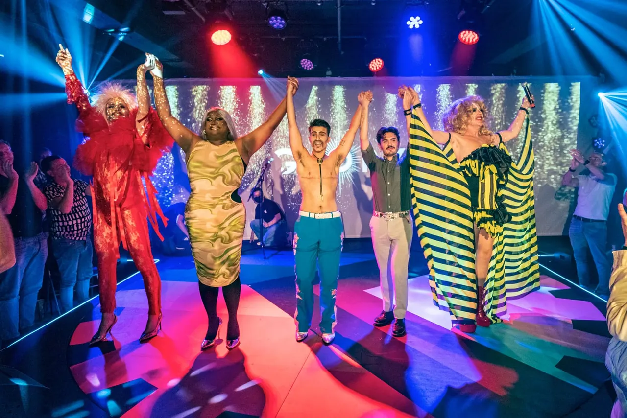Performers in colourful costumes dancing on a brightly lit stage with multi-coloured spotlights and glittering backdrop, audience members watching from the sides.