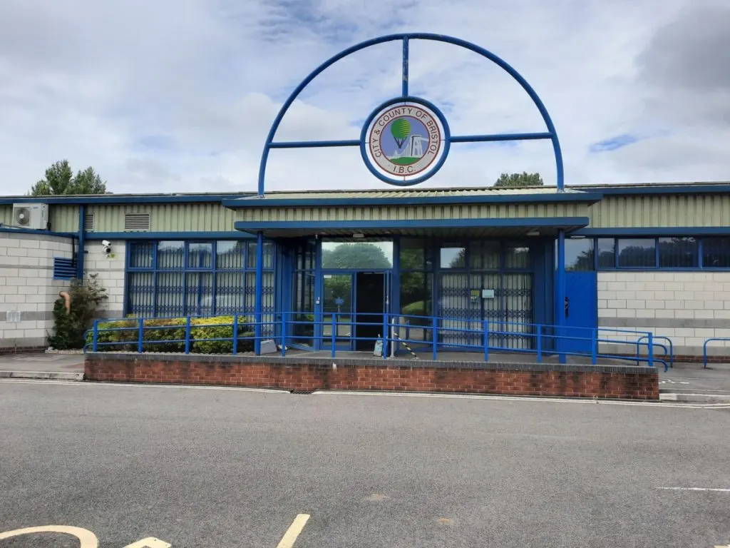 Modern sports pavilion with blue metal entrance canopy, white and corrugated metal walls, and a circular emblem above the doors.