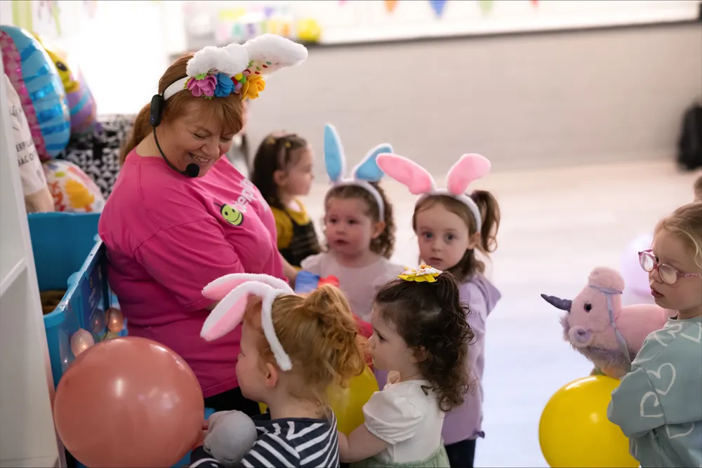 An adult in a pink shirt wearing a flower crown and bunny ears entertains a group of young children wearing bunny ear headbands at an indoor activity session.