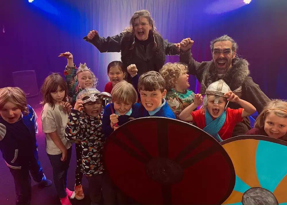 Two adults in costume entertain a group of excited children on a theatrical stage lit with purple and blue lighting.