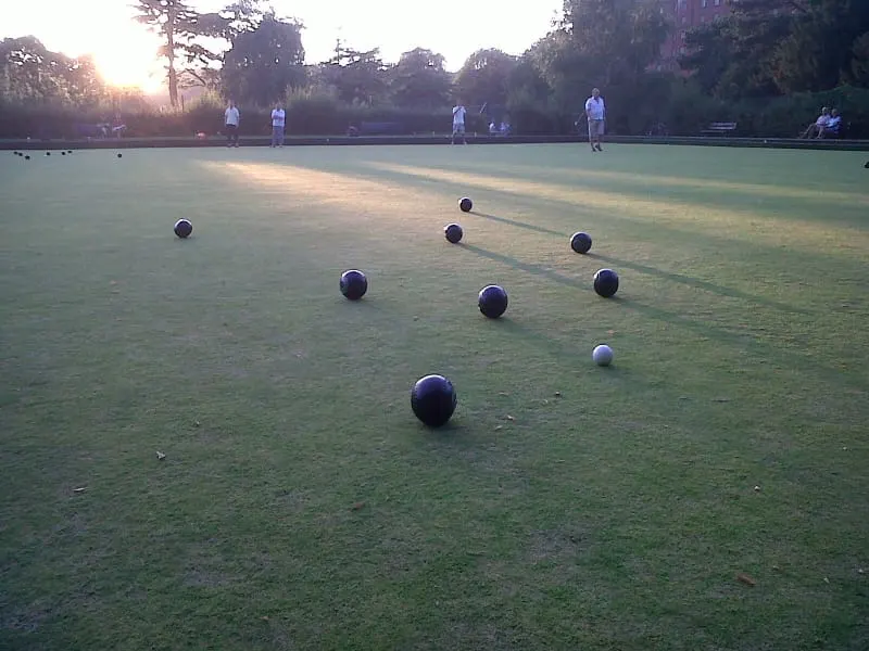 Bowls scattered across a green lawn during a match at sunset, with players standing in the background.