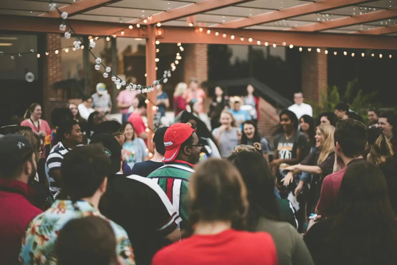 Large group of people gathered under a covered terrace with warm string lights and decorative hanging crystals for an evening event.
