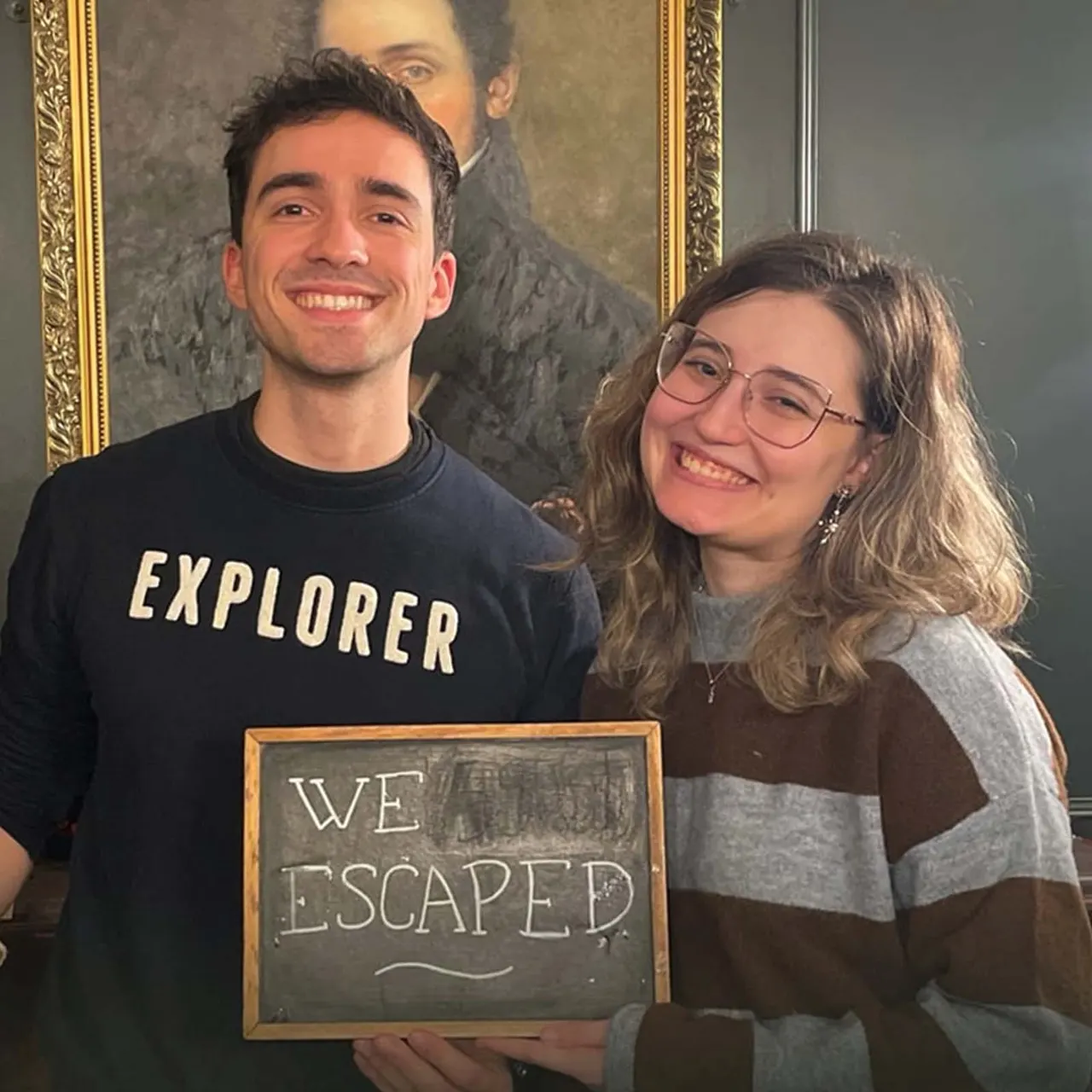 Two smiling visitors holding a "We Escaped" sign in front of a portrait painting at an escape room venue.