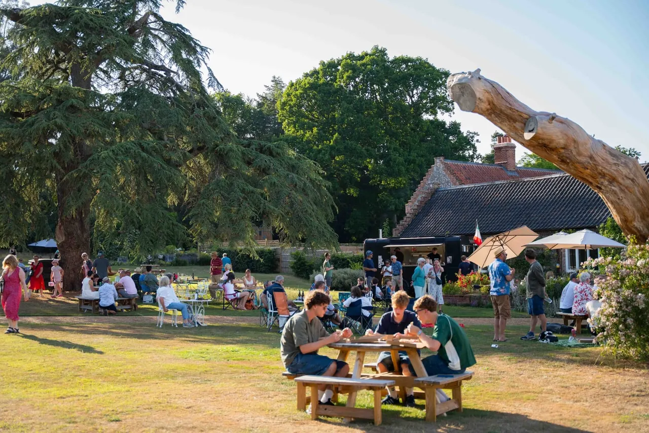 Visitors enjoy outdoor dining and leisure activities in a landscaped garden with picnic tables, mature trees, and a wooden sculpture, beside a brick manor house.