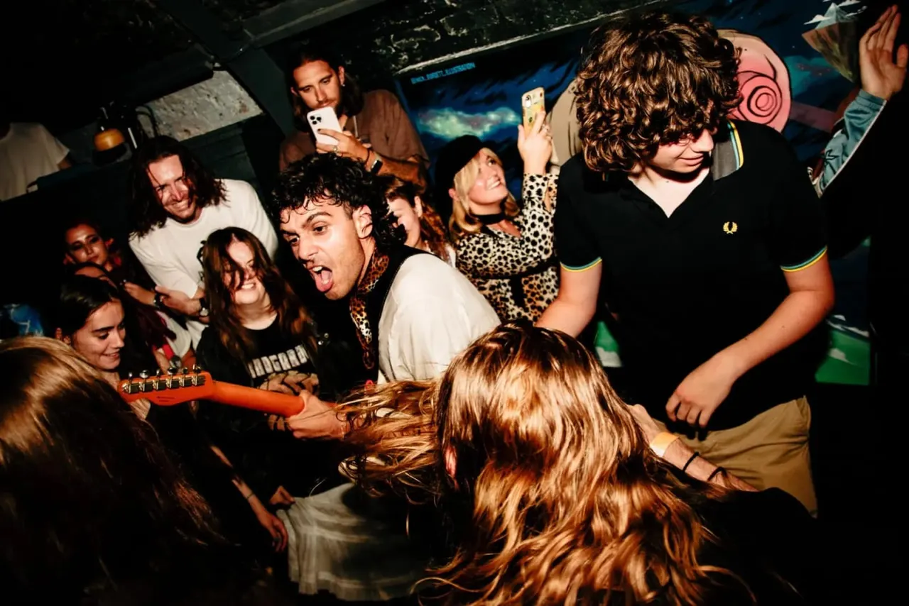 Group of young people dancing and socialising in a dimly lit nightclub, with someone holding a red electric guitar.