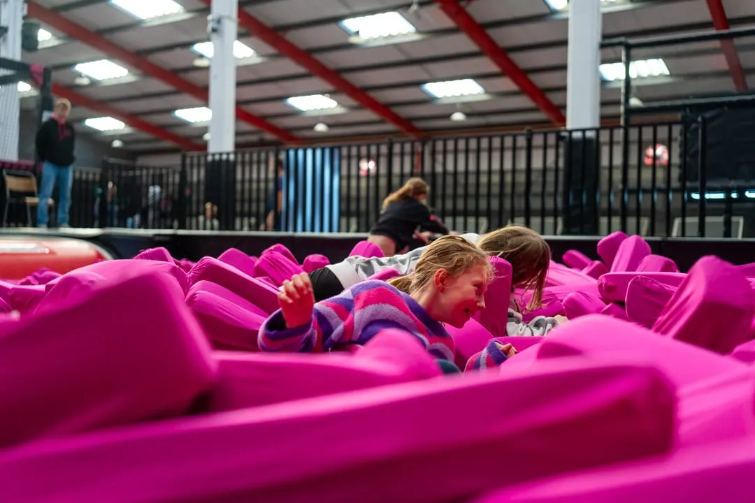 Children playing in a bright pink foam pit at an indoor leisure facility with metal railings and industrial ceiling.