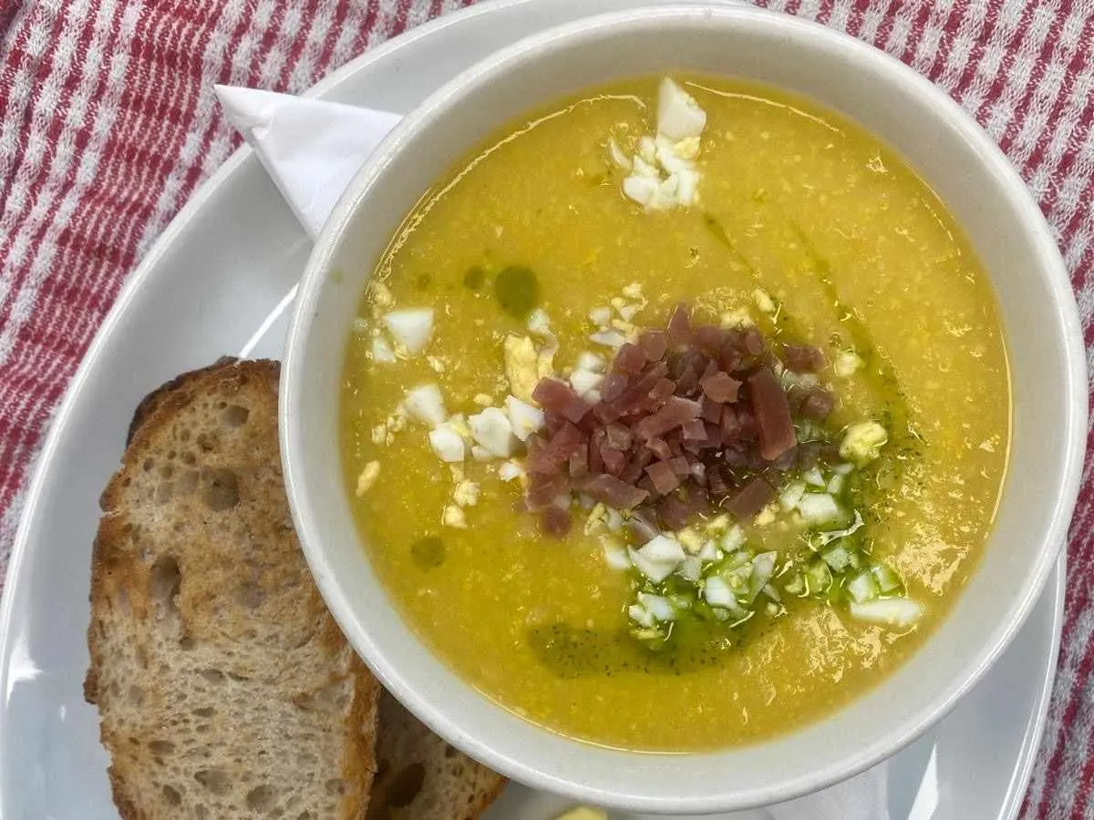 Bowl of yellow soup with ham, onion and greens, served with toasted bread on a striped cloth.