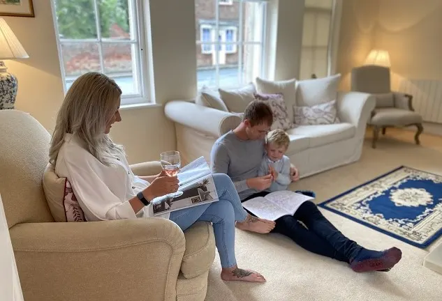 Woman reading in armchair whilst man and young child look at a book together on the floor in a bright, light-filled sitting room.