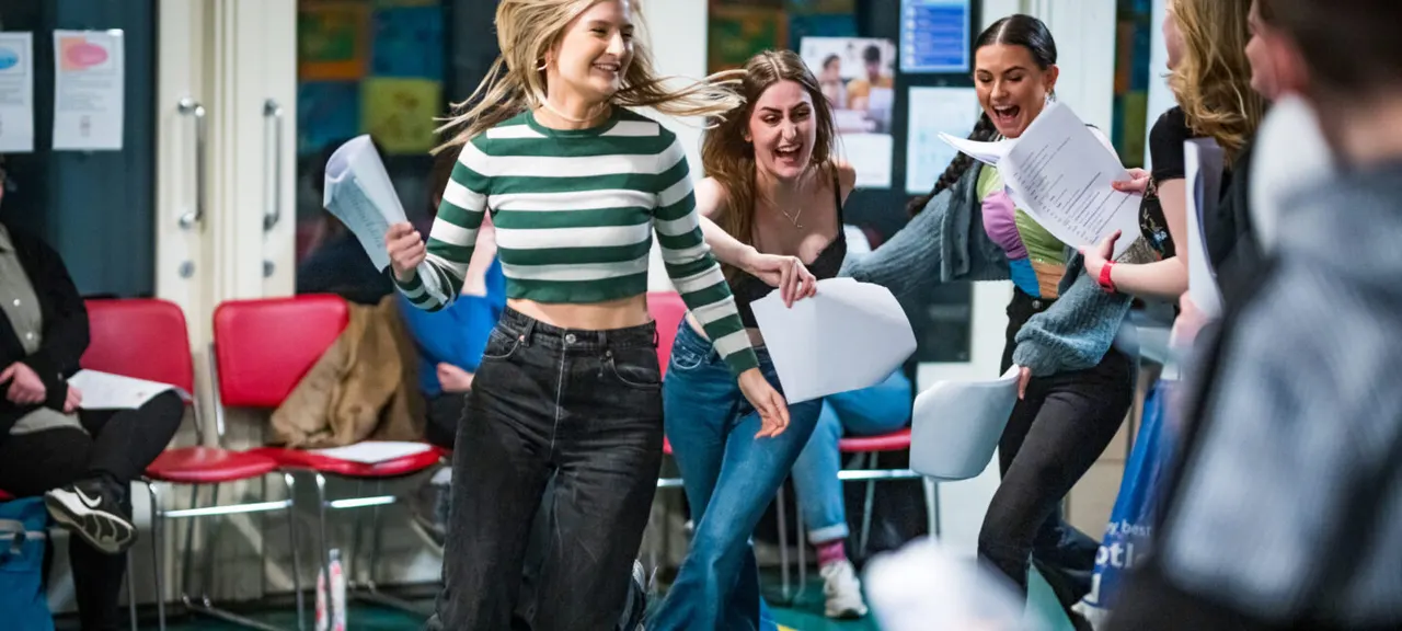 A group of young people laughing and dancing together in a modern indoor space with colourful chairs and notice boards.