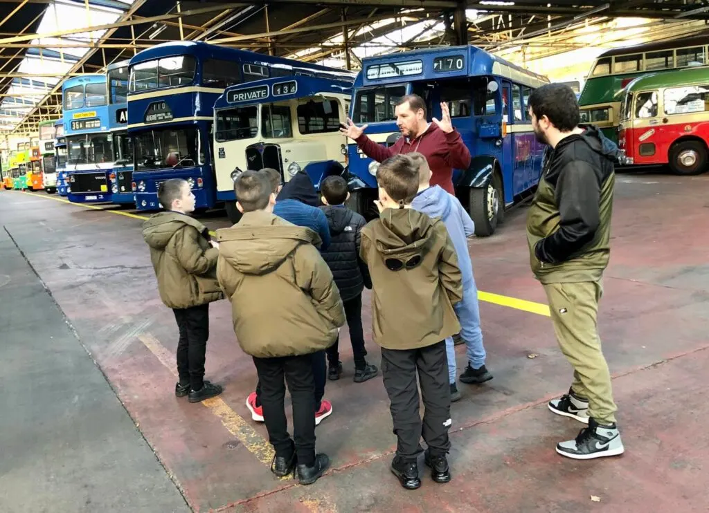 A group of schoolchildren and an adult guide stand in front of vintage double-decker buses in a garage depot.