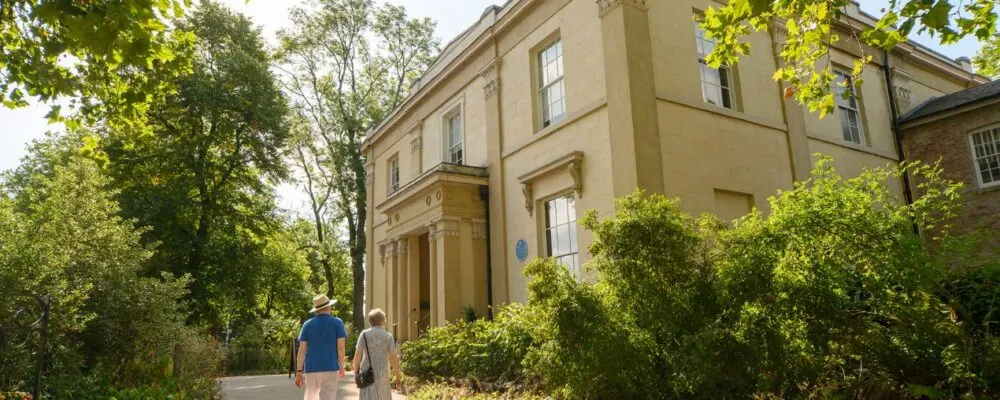 Two visitors walk along a tree-lined path towards a cream-coloured Georgian house on a sunny day.