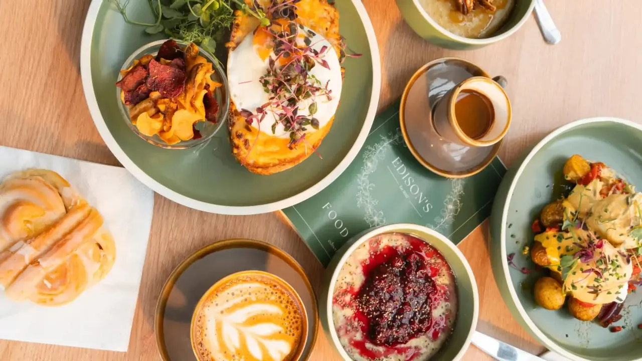 Overhead view of brunch dishes including roasted vegetables, toast, eggs, coffee with latte art, and a berry dessert on a wooden table.