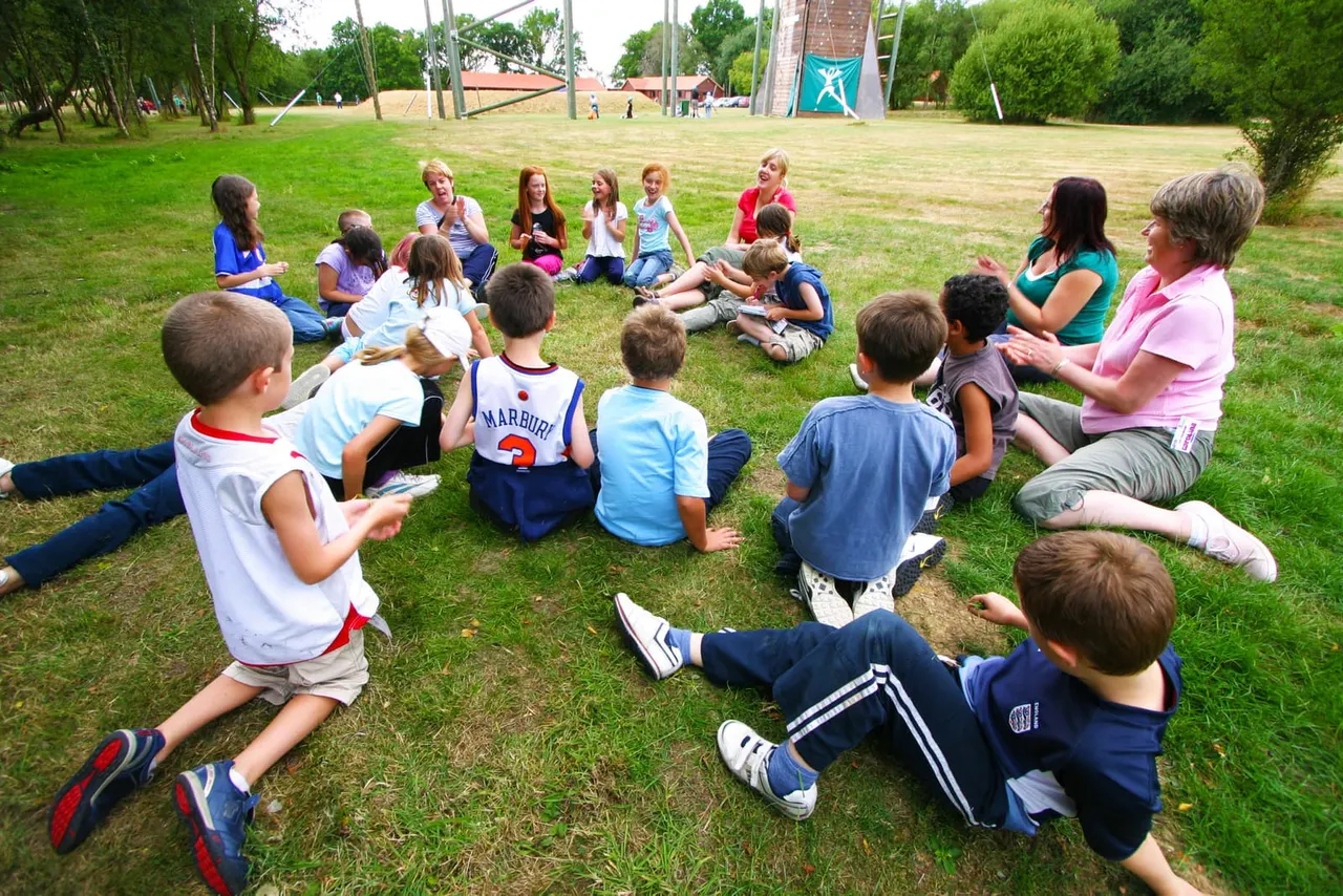 Large group of children and adults sitting in a circle on grass at an outdoor activity centre.