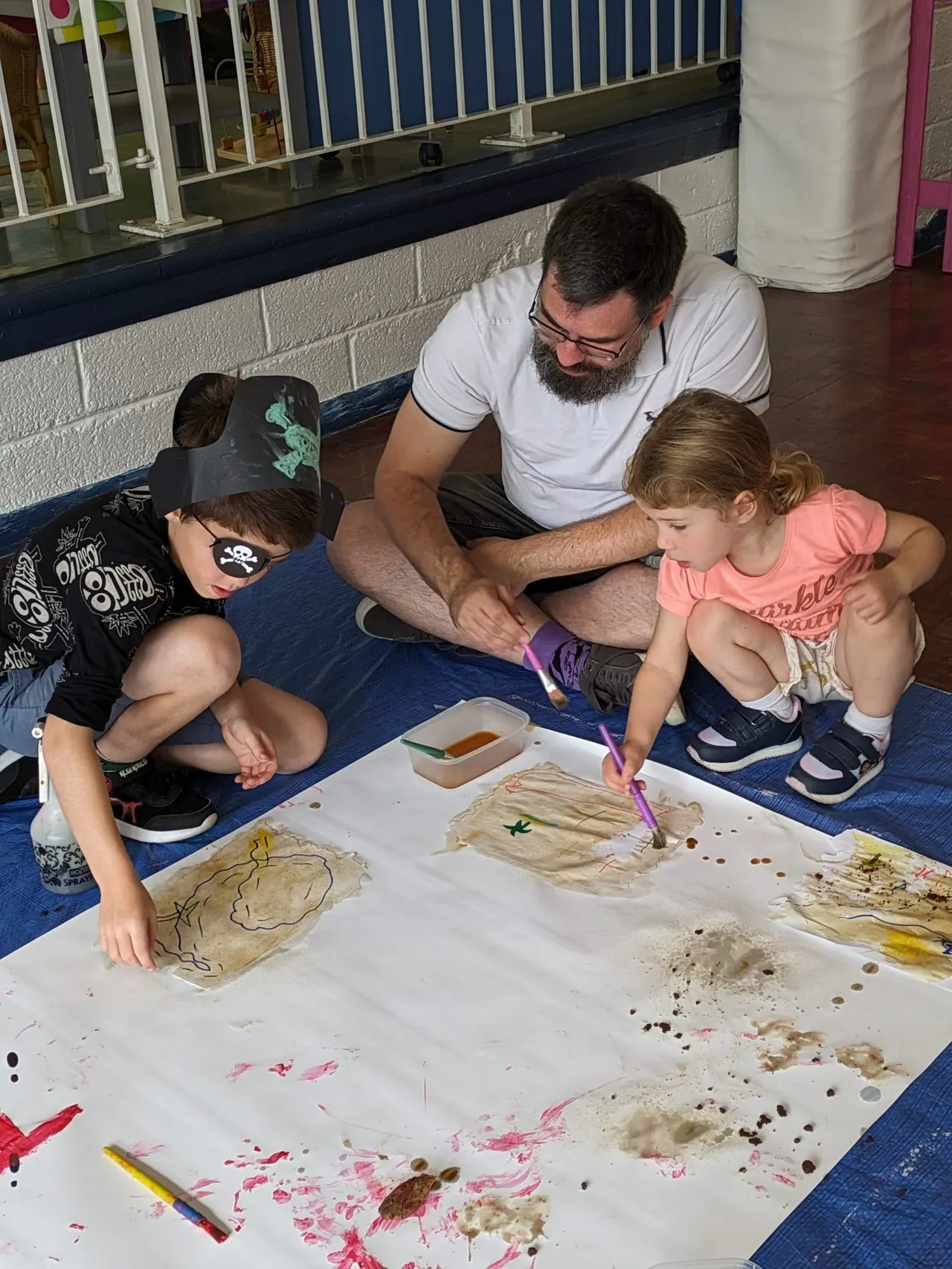 Adult and two children doing a messy painting activity together on the floor indoors.
