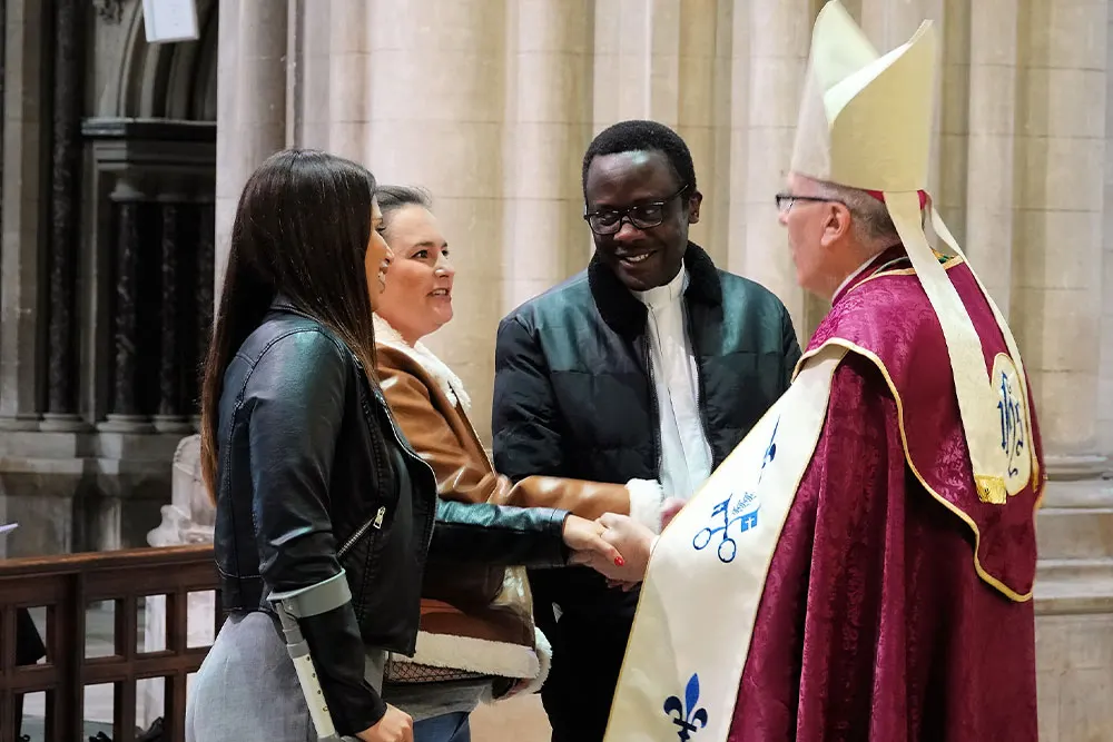 A bishop in ceremonial vestments shakes hands with three visitors inside a cathedral with stone columns.