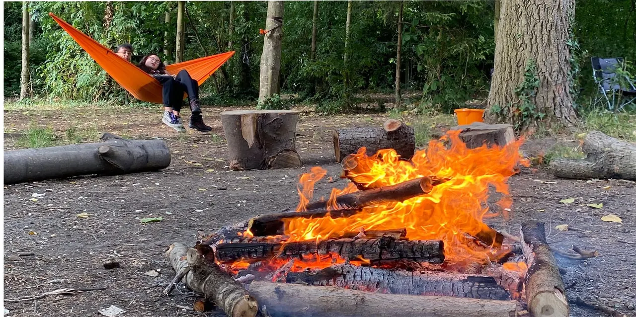 Two people relaxing in an orange hammock beside a blazing campfire in a woodland clearing.