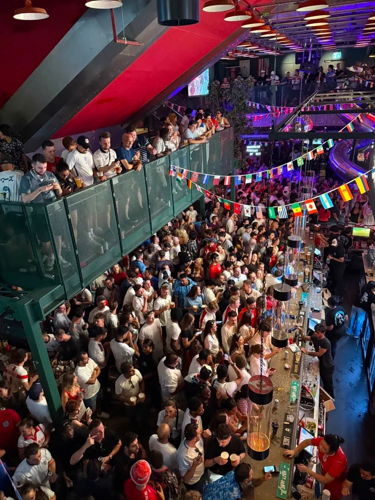 Crowded indoor venue with people standing and socialising on multiple levels, decorated with colourful bunting and lit by purple and red lighting.