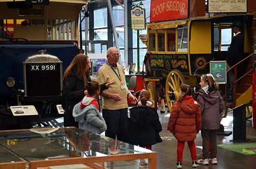 A museum guide talks to families with children near vintage buses and vehicles on display.