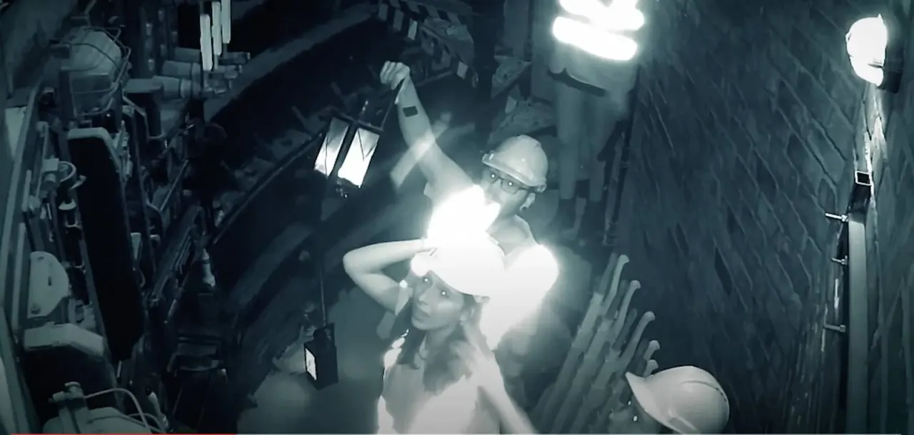 Two people wearing hard hats explore a dark industrial tunnel lit by handheld torches and overhead lights.