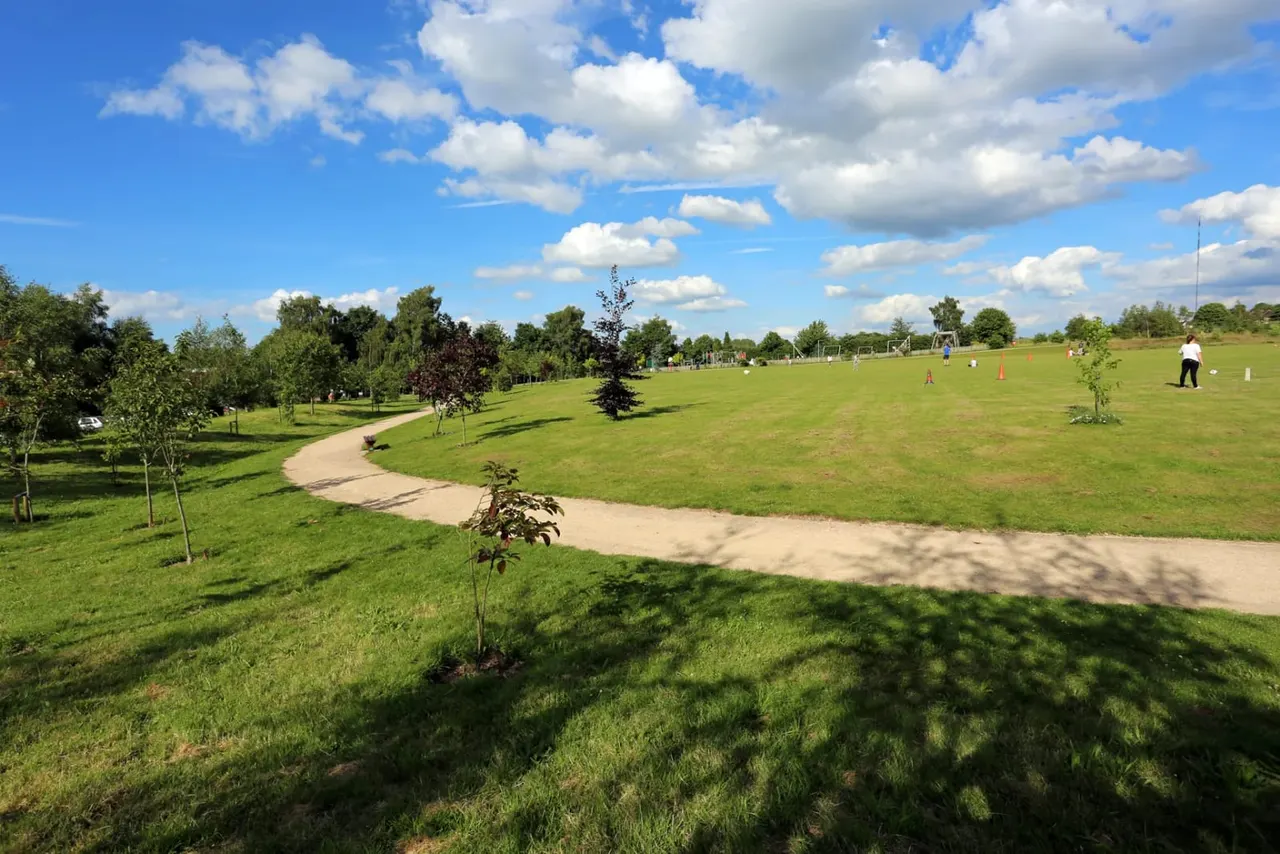 A spacious green park with curved paths, young trees, and people enjoying recreational activities under a blue cloudy sky.