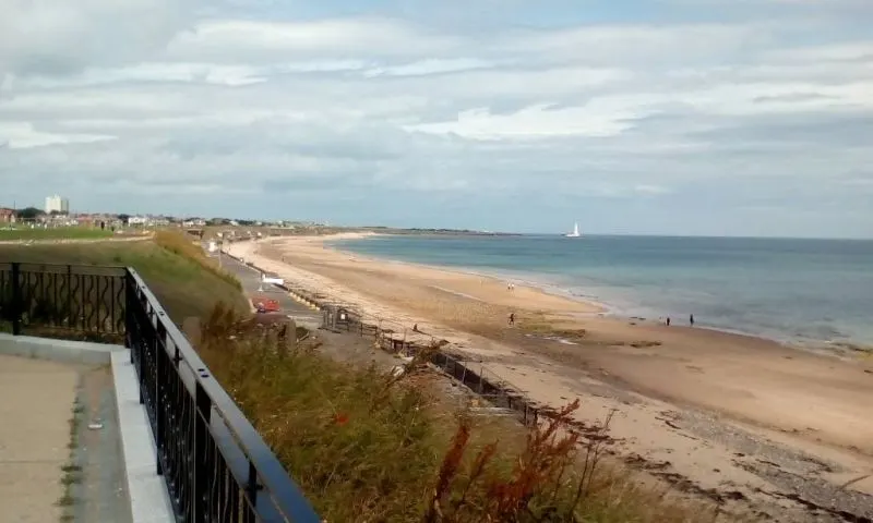 Wide sandy beach with turquoise sea, viewed from clifftop promenade railing, coastal town in distance.