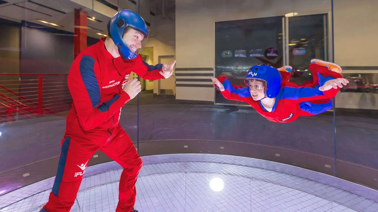 An instructor in red suit and blue helmet guides a child in matching gear during indoor skydiving in a wind tunnel chamber.