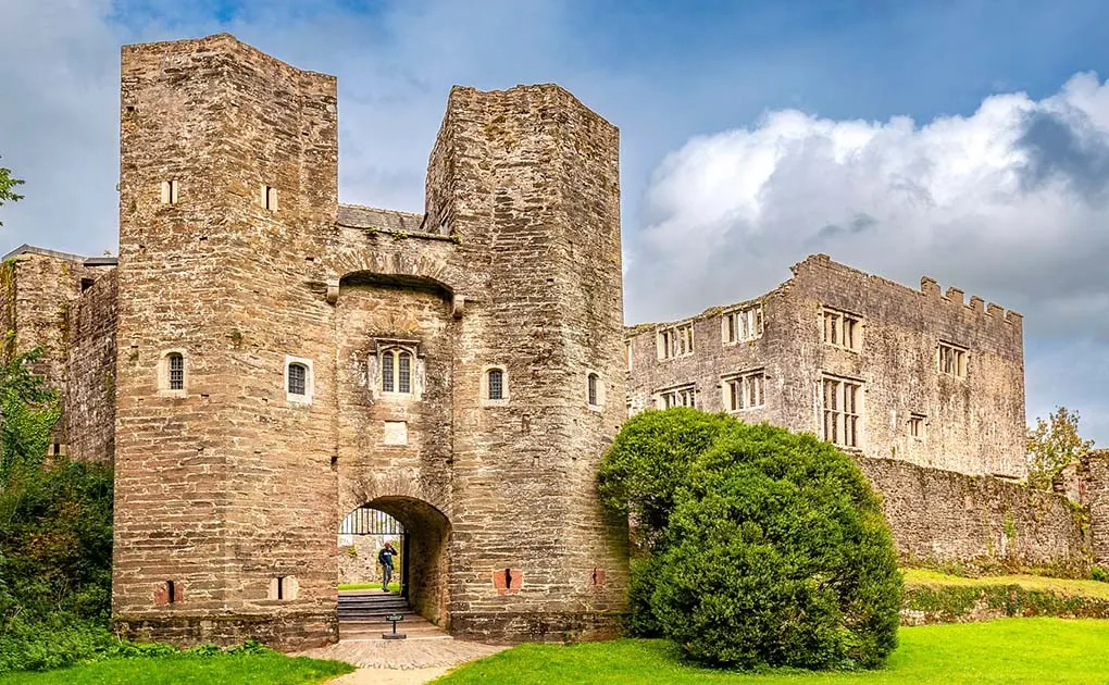 Medieval stone castle ruins with twin towers and gatehouse, surrounded by green grounds and ivy-covered walls under blue sky.