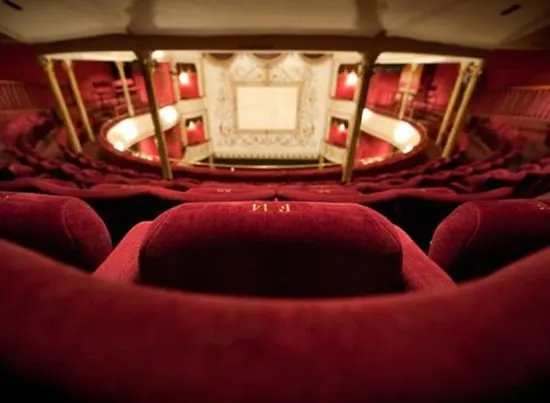 Red velvet theatre seats facing an ornate stage with multiple levels and gold-framed boxes in an empty auditorium.