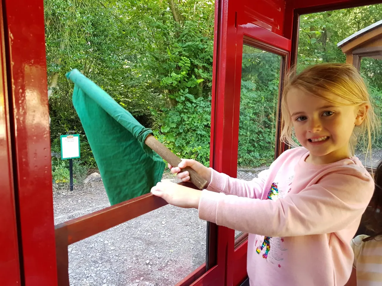 Young child in pink top feeding a green fabric bird puppet through a red-framed window at a leisure venue.
