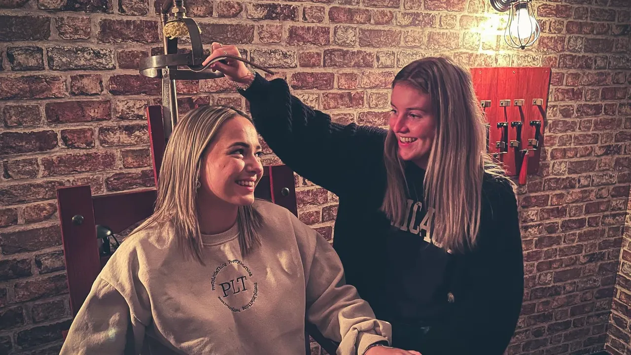 Two young women laugh together in a brick-walled escape room with period props and red equipment on the wall.