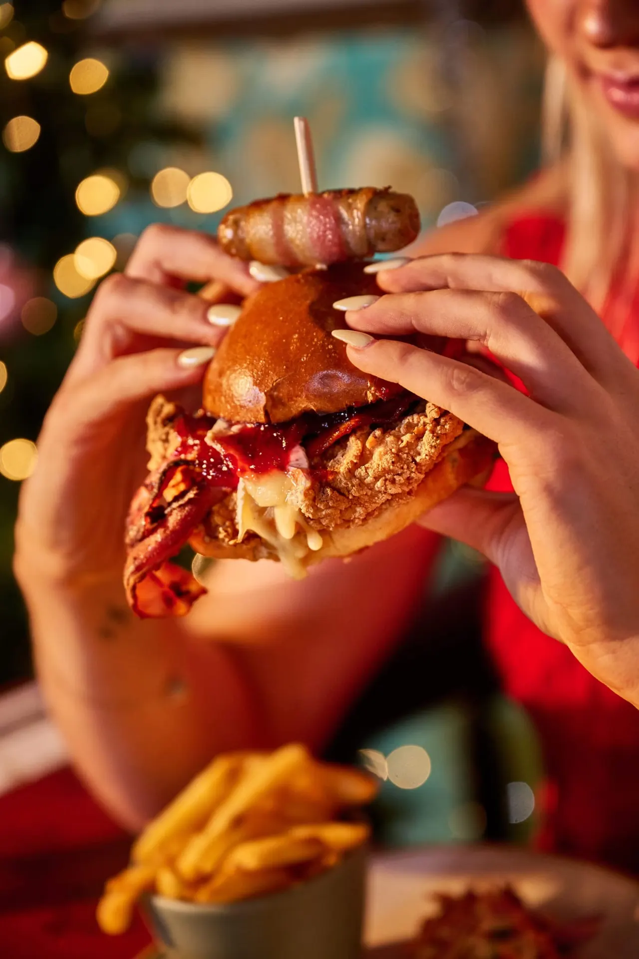 Person in red holding a loaded burger with fried chicken, sauce and a skewer, with festive lights and chips nearby.