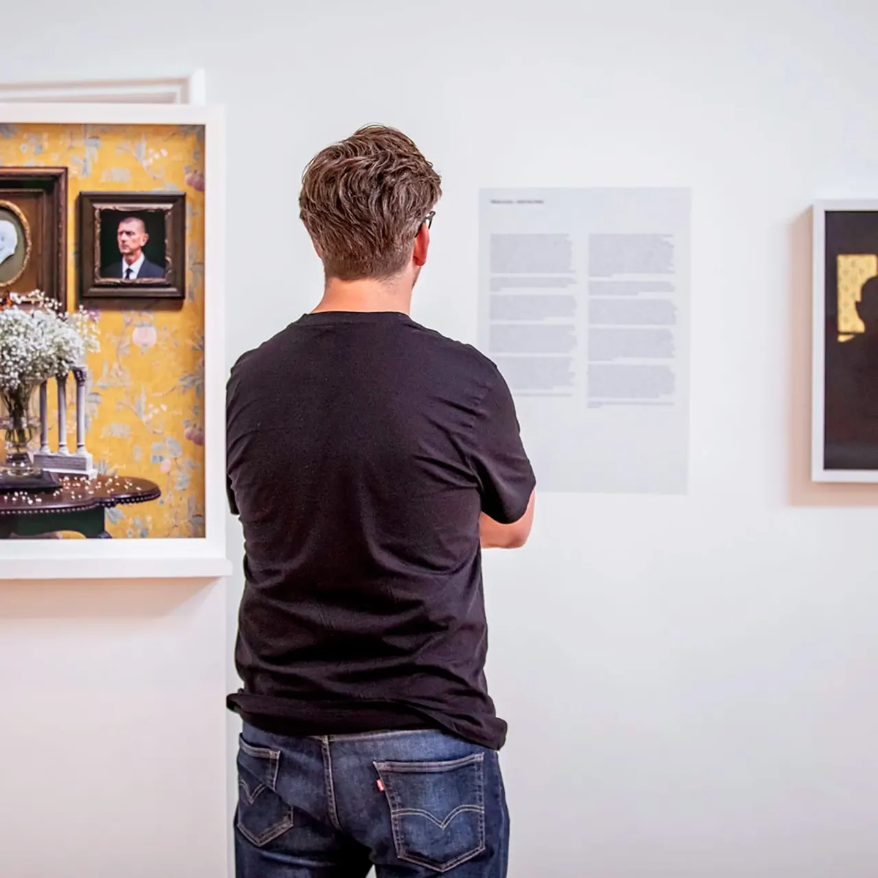 A visitor viewed from behind examines framed artworks and text panels displayed on white gallery walls.