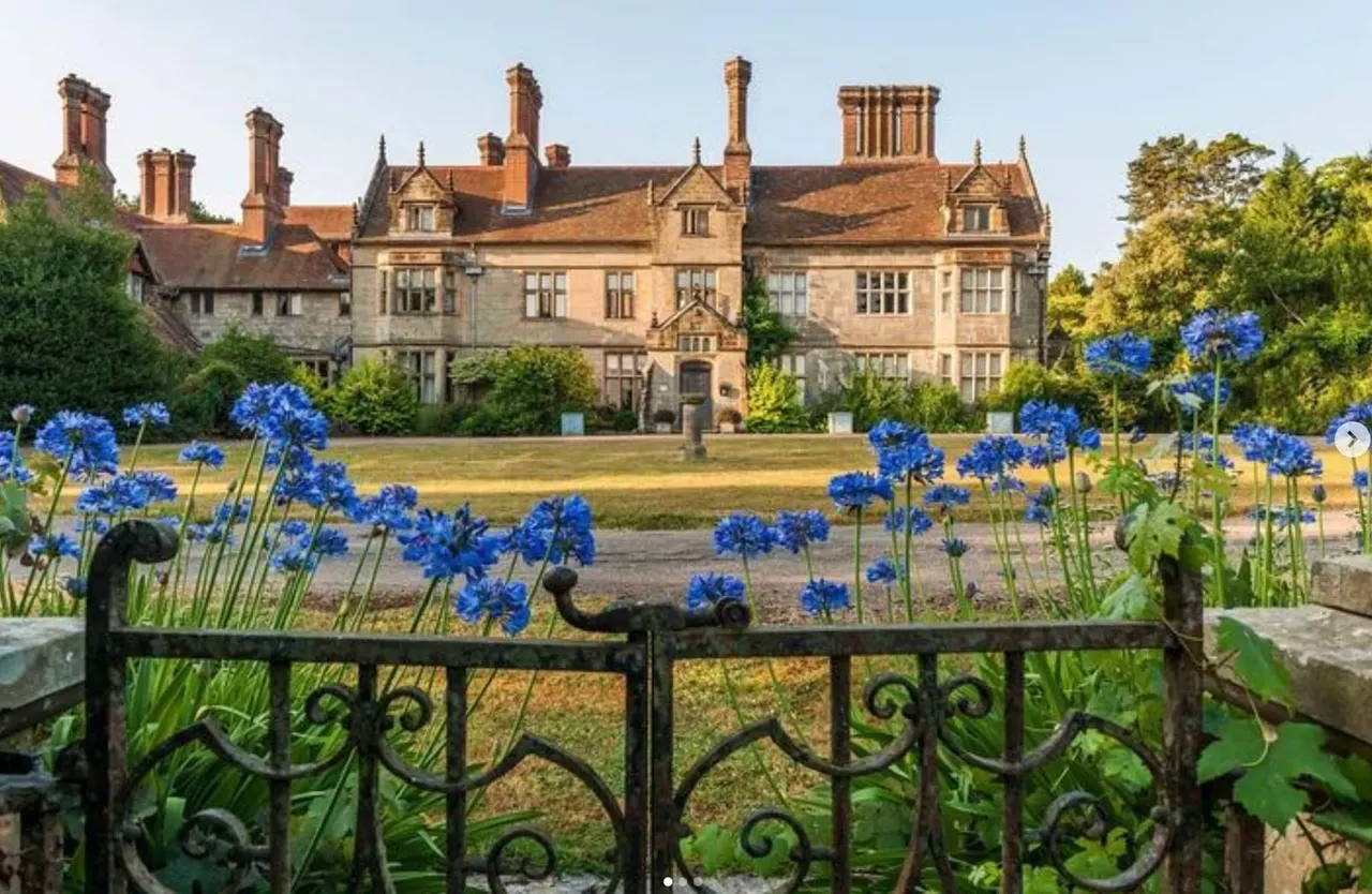 Large stone country house with ornamental chimneys viewed across formal gardens with blue flowers and an ornate black iron fence in the foreground.