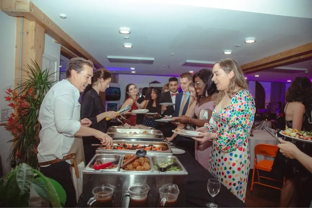 Guests at an indoor event serving themselves food from a buffet line under purple mood lighting.