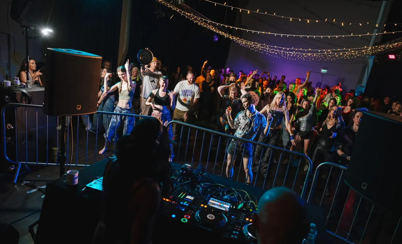 Large crowd of people dancing and celebrating at a nightclub event with colourful lighting and string lights overhead.