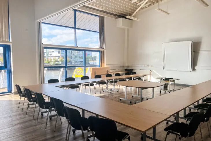 Bright, modern training room with wooden tables, black chairs, and large windows overlooking water and buildings.