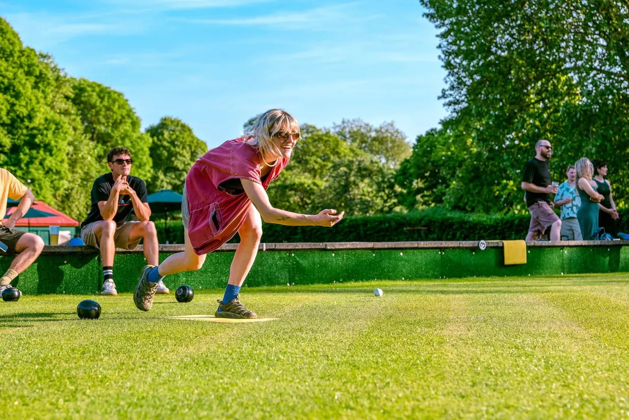A young person in a burgundy shirt plays lawn bowls on a green, with spectators watching from the sidelines on a sunny day.