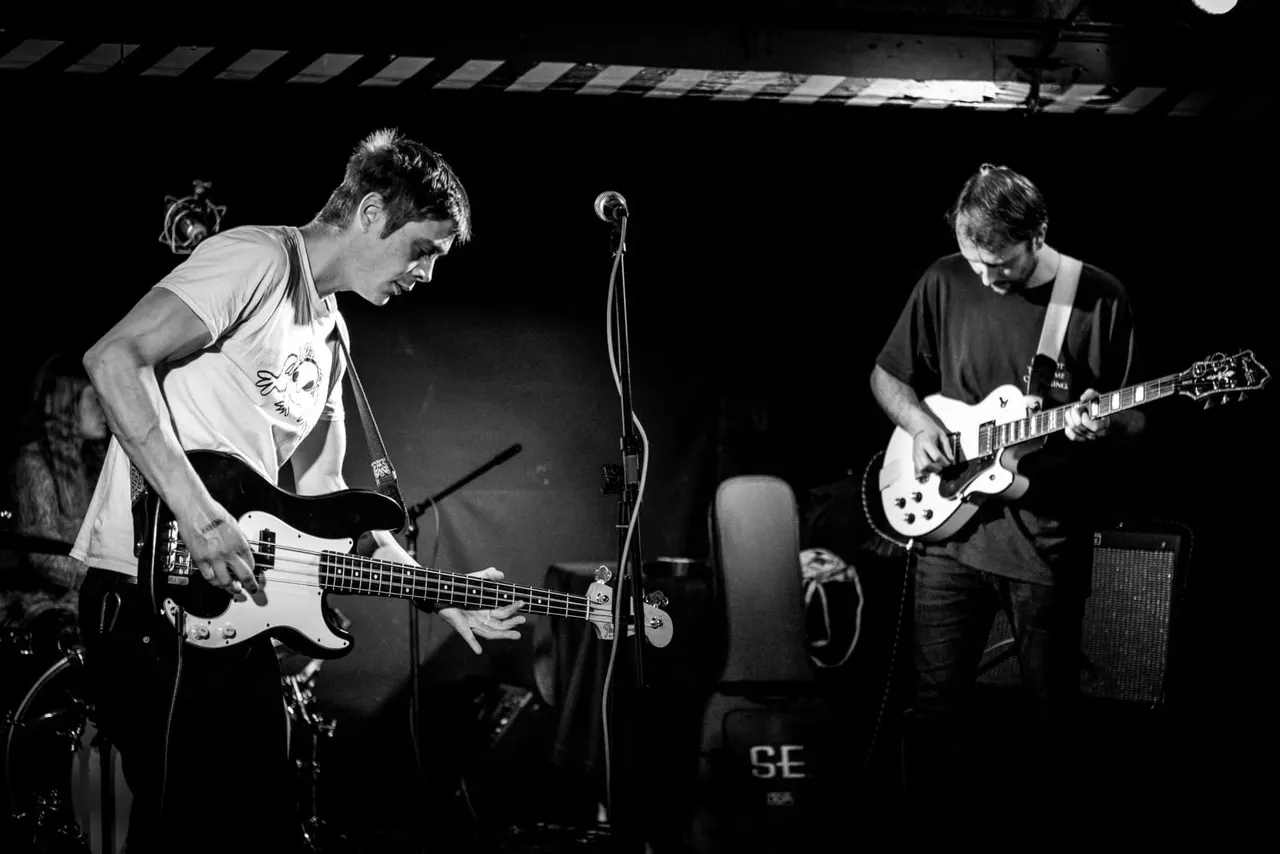 Two musicians performing on stage with electric guitars under stage lighting in a black and white photograph.