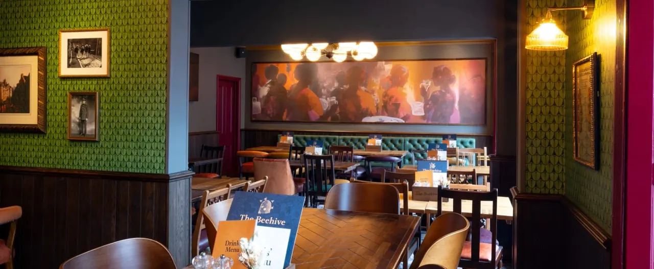 Interior of a pub with wooden tables and chairs, green patterned wallpaper, framed photographs, and a screen showing people dancing.