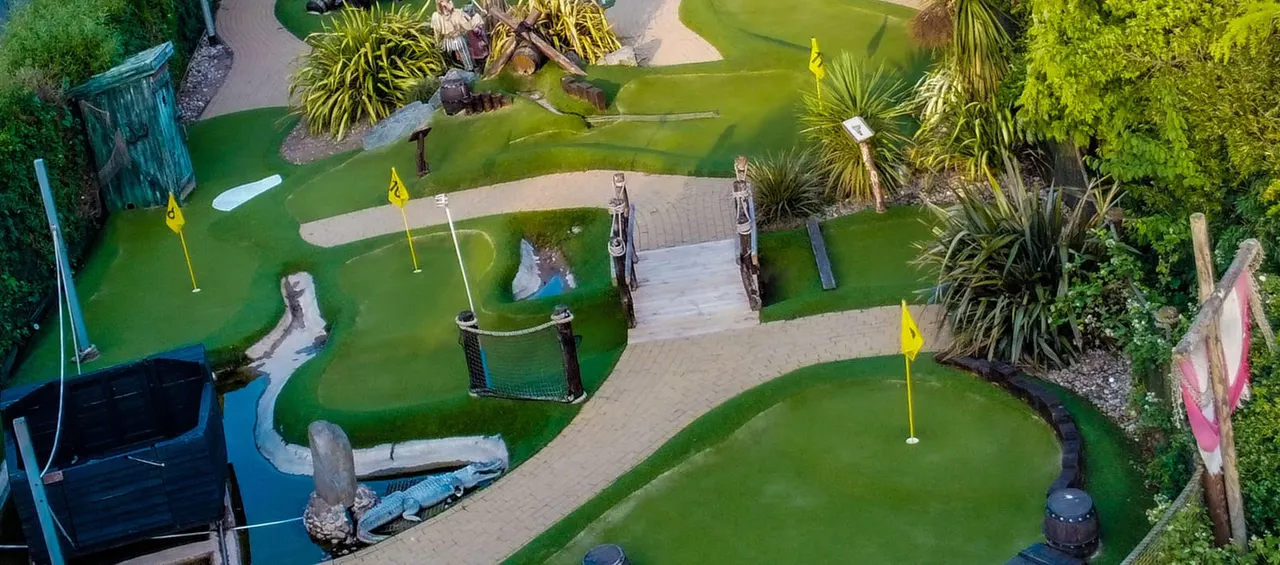 Aerial view of a themed mini golf course with green fairways, sand bunkers, flags, and pirate-themed obstacles including a crocodile sculpture and wooden bridge.
