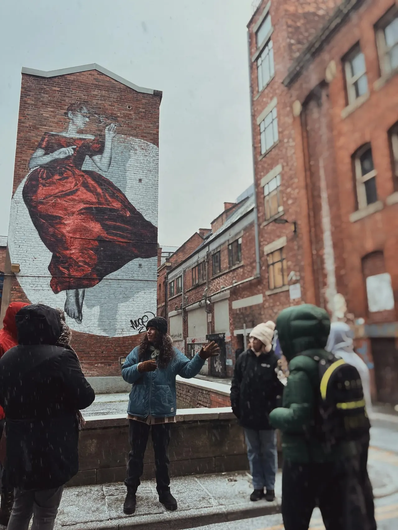Visitors on a guided street art tour view a large mural of a figure in a red dress on a brick building in an urban setting.