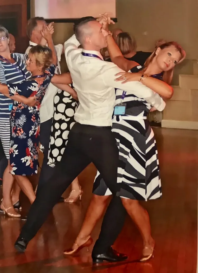 Adults dancing together on a wooden dance floor at an indoor event, with spectators watching in the background.