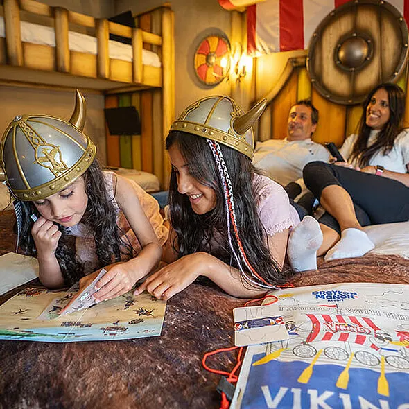 Two children wearing Viking helmets play with activity sheets on a bed while adults watch in a themed hotel room with Viking decorations.