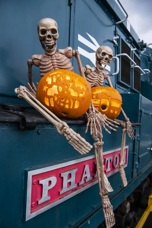Skeleton figures holding illuminated jack-o'-lanterns mounted on the side of a heritage railway vehicle.