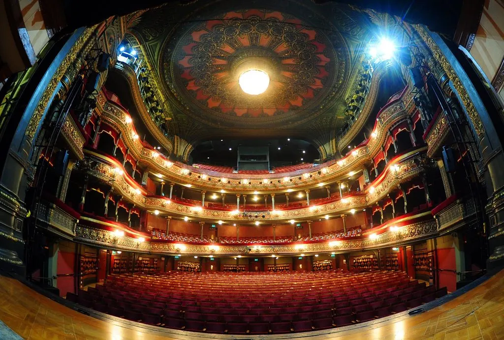 Interior view of an ornate theatre auditorium with multiple tiers of red velvet seats, gilded balconies, and an illuminated decorative ceiling dome.