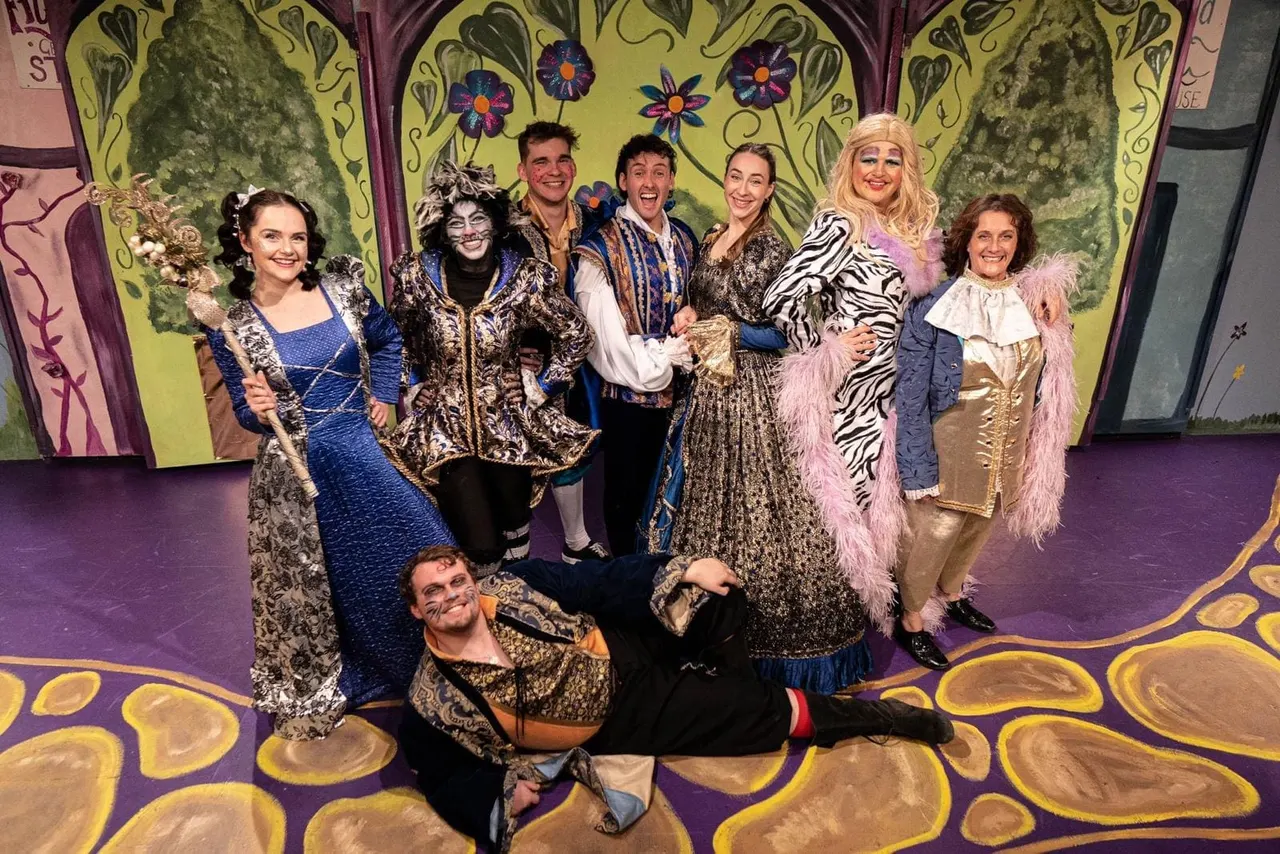 Cast members in colourful fairytale costumes pose together on a purple stage set with ornate yellow and green painted backdrop.